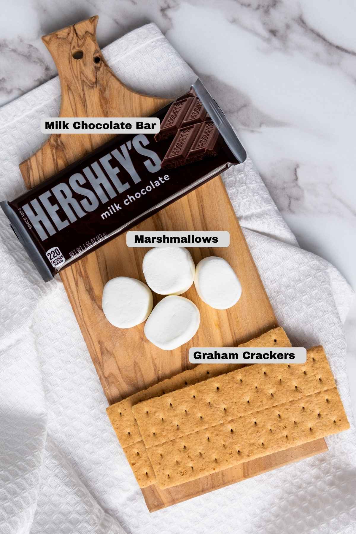 Ingredients for making indoor s'mores to include Hershey's milk chocolate bar, marshmallows, and graham crackers labeled and arranged on a wooden board.