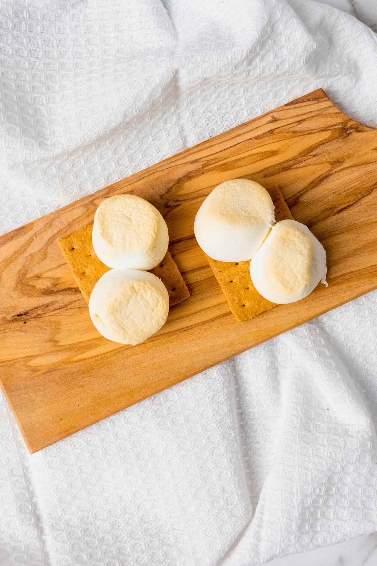 Two graham cracker halves topped with toasted marshmallows on a wooden board over a white textured cloth.