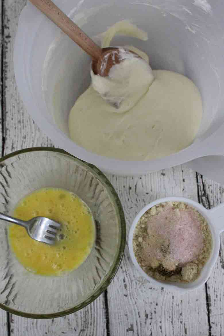 Mixing bowls with melted cheese dough, beaten egg, and almond flour mixture on a rustic white wood surface.