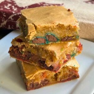 Close up of 3 stacked brown butter M&M Blondie Bars on a white plate, showing a golden crust and colorful candy pieces inside.