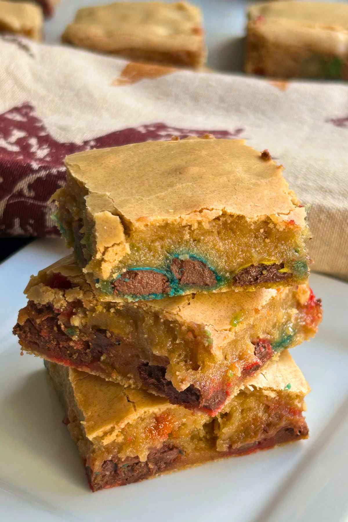 Close up of 3 stacked brown butter M&M Blondie Bars on a white plate, showing a golden crust and colorful candy pieces inside.