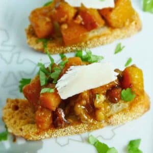 Close-up of butternut squash crostini topped with caramelized cubes of squash, chopped nuts, and a thin slice of cheese on toasted bread, garnished with fresh cilantro.