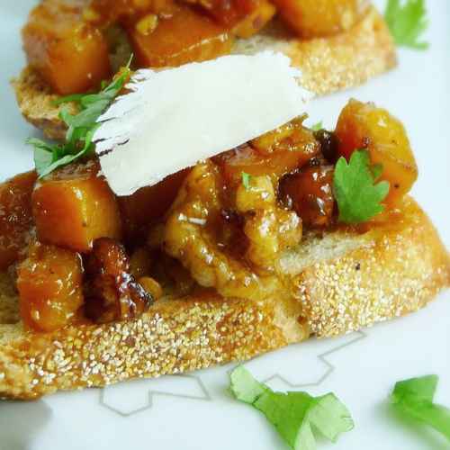 Close-up of butternut squash crostini topped with caramelized cubes of squash, chopped nuts, and a thin slice of cheese on toasted bread, garnished with fresh cilantro.