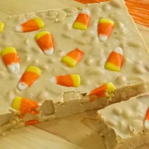 A close up of creamy beige fudge topped with candy corn pieces, sowing a smooth surface with a few nuts mixed in and one piece cut from the slab.