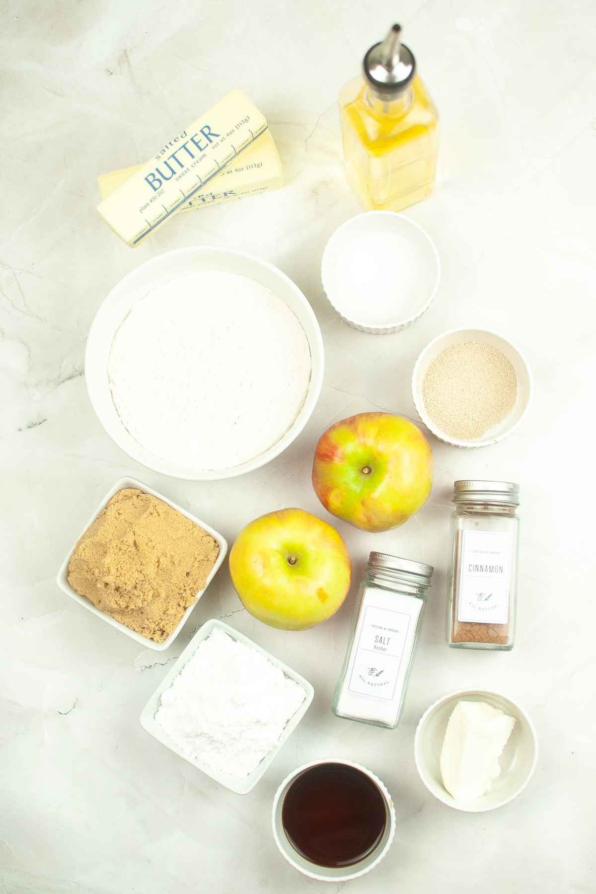 Flat lay of baking ingredients including apples, flour, brown sugar, powdered sugar, butter, oil, cinnamon, salt, yeast, and cream cheese arranged on a marble surface.