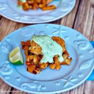 A plated dish featuring a crispy piece of chicken served over sweet potato waffle fries, topped with a creamy green sauce and garnished with a lime wedge.