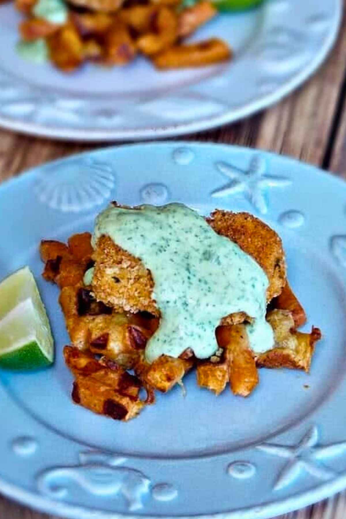 A plated dish featuring a crispy piece of chicken served over sweet potato waffle fries, topped with a creamy green sauce and garnished with a lime wedge.