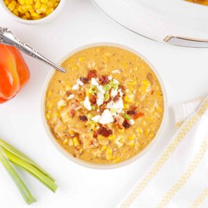 Overhead view of a bowl of chipotle corn chowder topped with bacon, green onions, and crumbled cheese, surrounded by corn and a red bell pepper.