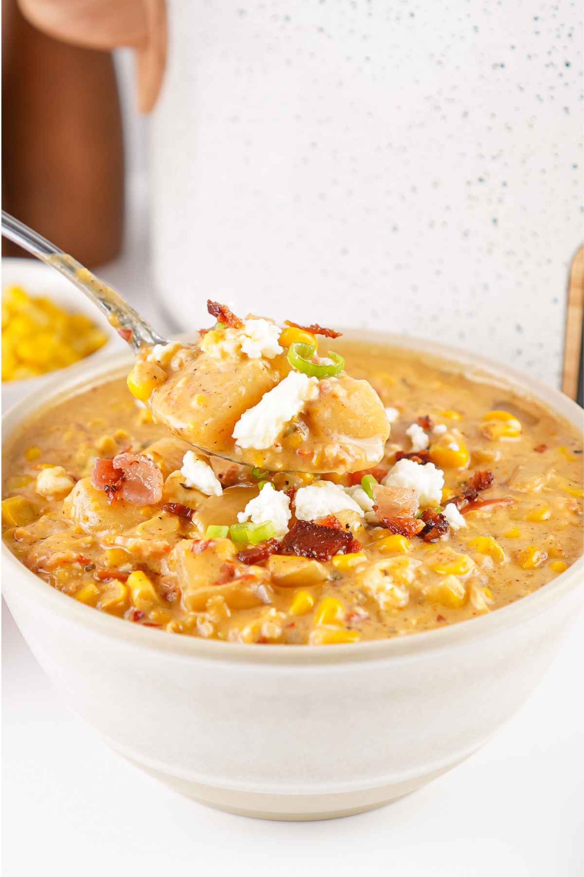A spoonful of creamy chipotle corn chowder topped with bacon, green onions, and cheese held above a bowl filled with the same hearty soup.