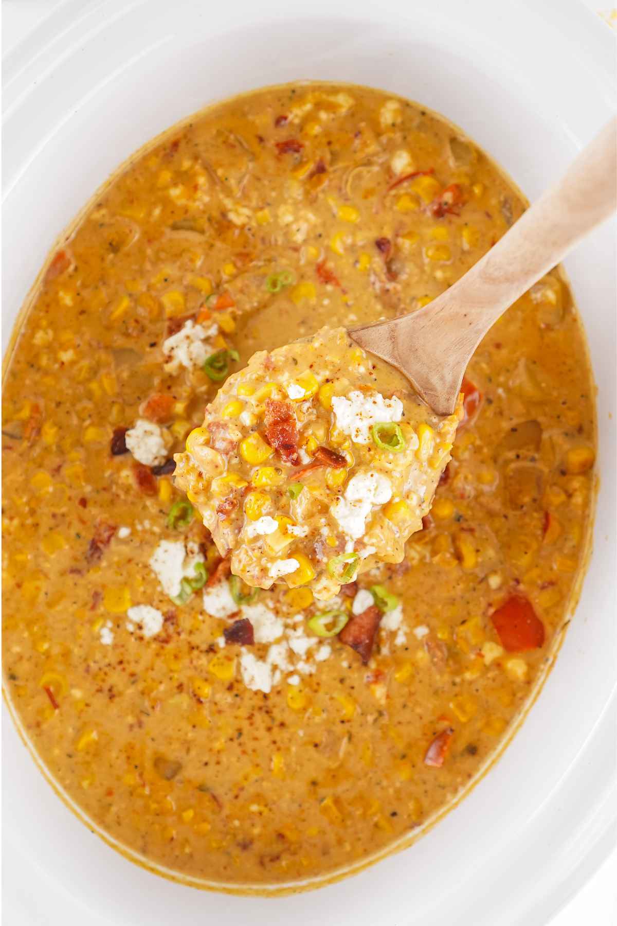 Overhead view of slow cooker corn chowder in a white pot, with a wooden spoon lifting a serving filled with corn, bacon, cheese, and peppers.