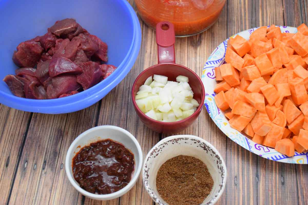 Ingredients for Chipotle Venison stew arranged on a wooden surface, including cubed venison in a blue bowl topped with a generous amount of dark seasoning blend.