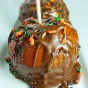 Close up of a caramel apple coated in melted chocolate and topped with colorful fall-themed sprinkles, served on a light plate with a white stick inserted on top.