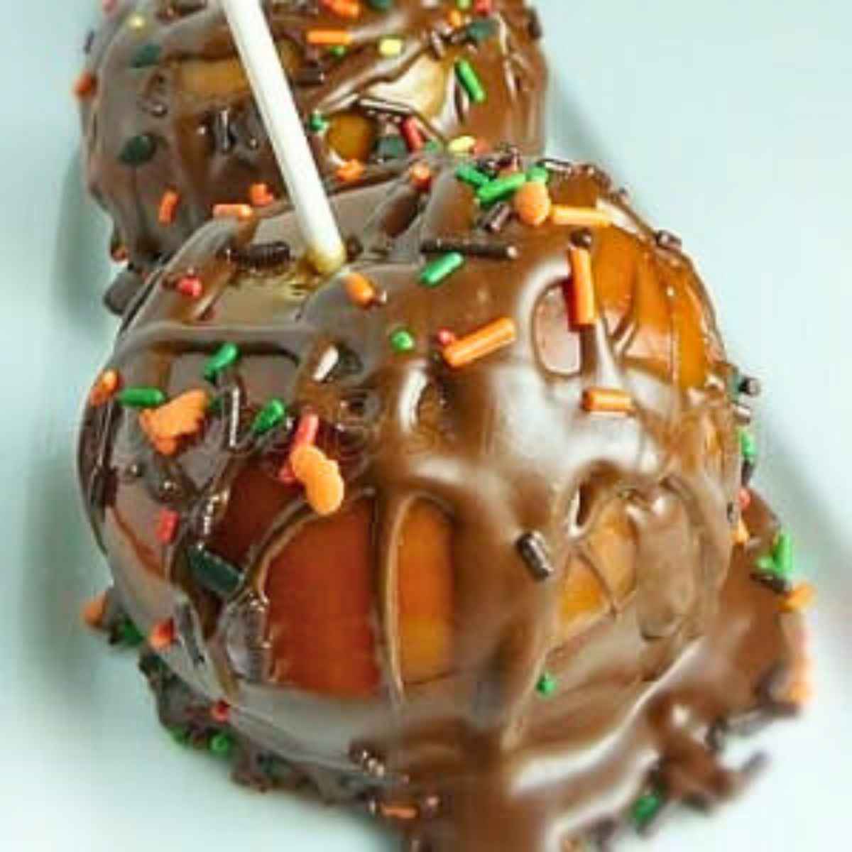 Close up of chocolate dipped caramel apples topped with colorful fall-themed sprinkles, served on a light plate with a white stick inserted on top.