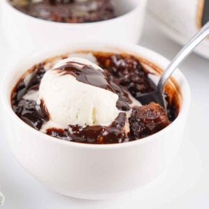 A white bowl filled with warm chocolate cobbler topped with melting vanlla ice cream and chocolate sauce, with another serving visible in the background.