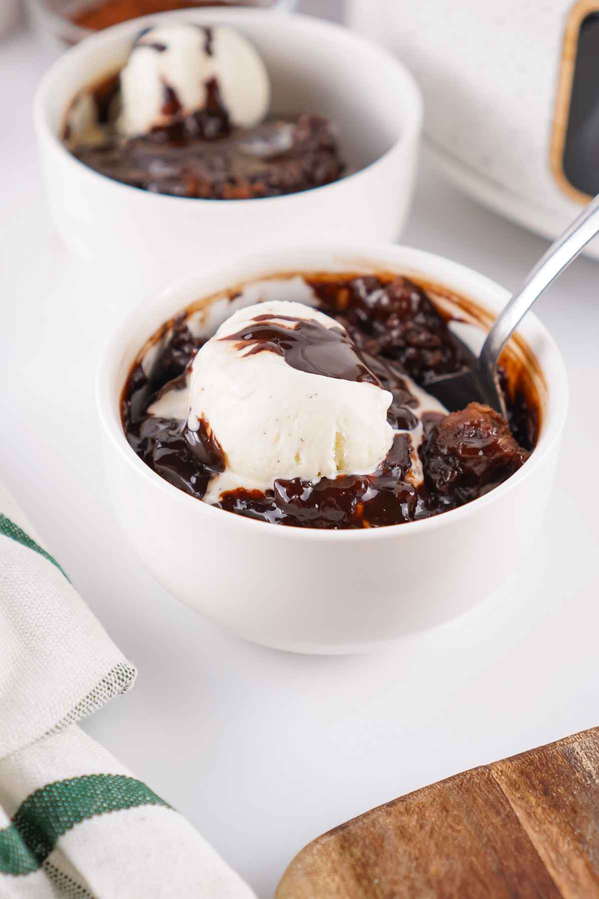 Two white bowls of chocolate cobbler topped with scoops of vanilla ice cream, placed beside a slow cooker and a small bowl of cocoa powder.