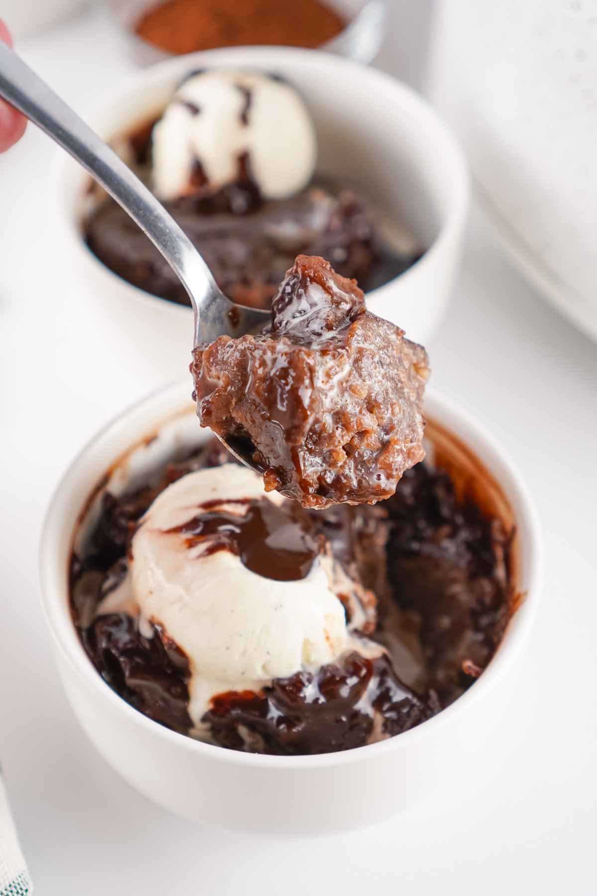 A spoonful of gooey chocolate cobbler being lifted from a white bowl topped with a scoop of vanilla ice cream drizzled with chocolate sauce.