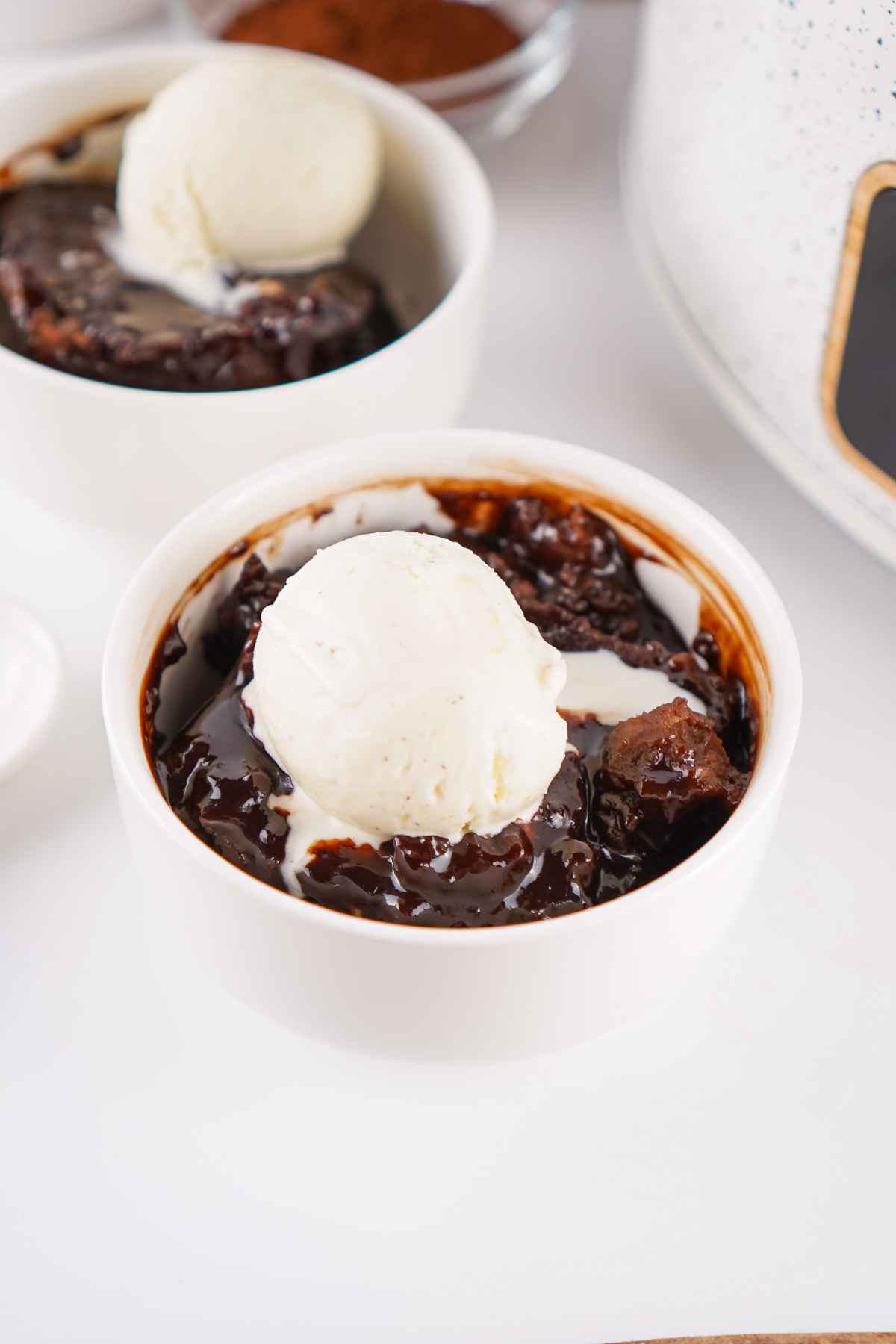 Two white bowls of chocolate cobbler topped with scoops of vanilla ice cream, placed beside a slow cooker and a small bowl of cocoa powder.