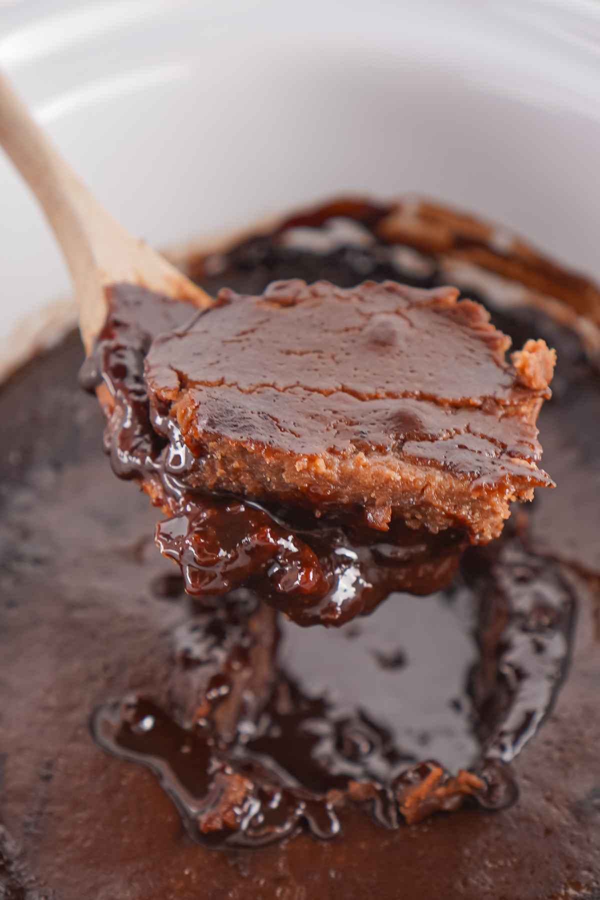 A wooden spoon lifting a serving of chocolate cobbler from the slow cooker, showing its moist cake layer and rich chocolate sauce underneath.