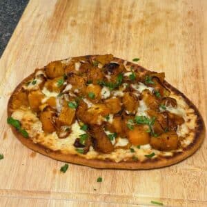 Whole flatbread pizza topped with golden roasted butternut squash, caramelized onions, melted cheese, and sprinkled herbs on a wooden board.