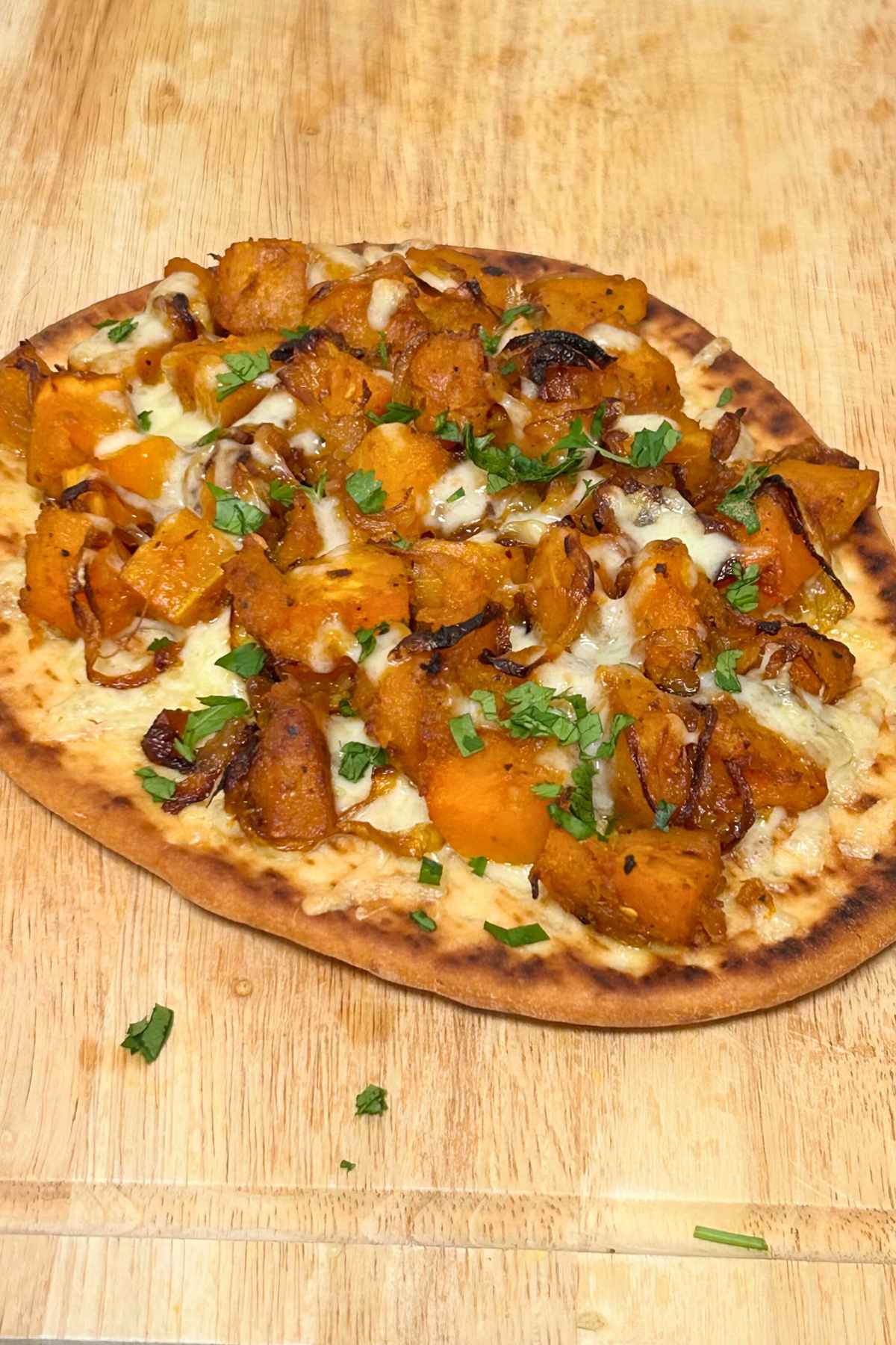 Oval flatbread covered with chunks of curried butternut squash, caramelized onions, melted cheese, and chopped cilantro resting on a wooden board.