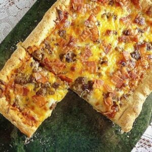 A rectangular deep dish bacon cheeseburger pizza with a thick crust, melted cheese, ground beef, and bacon pieces with one slice removed to show the interior.