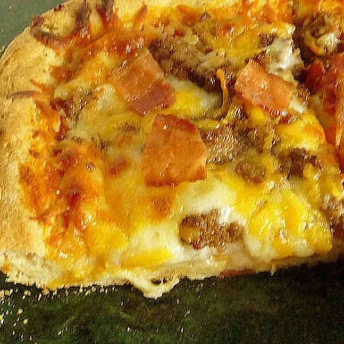 A close-up view of a deep-dish bacon cheeseburger pizza slice, showing gooey melted cheese, ground beef, and crispy bacon on a thick golden crust.