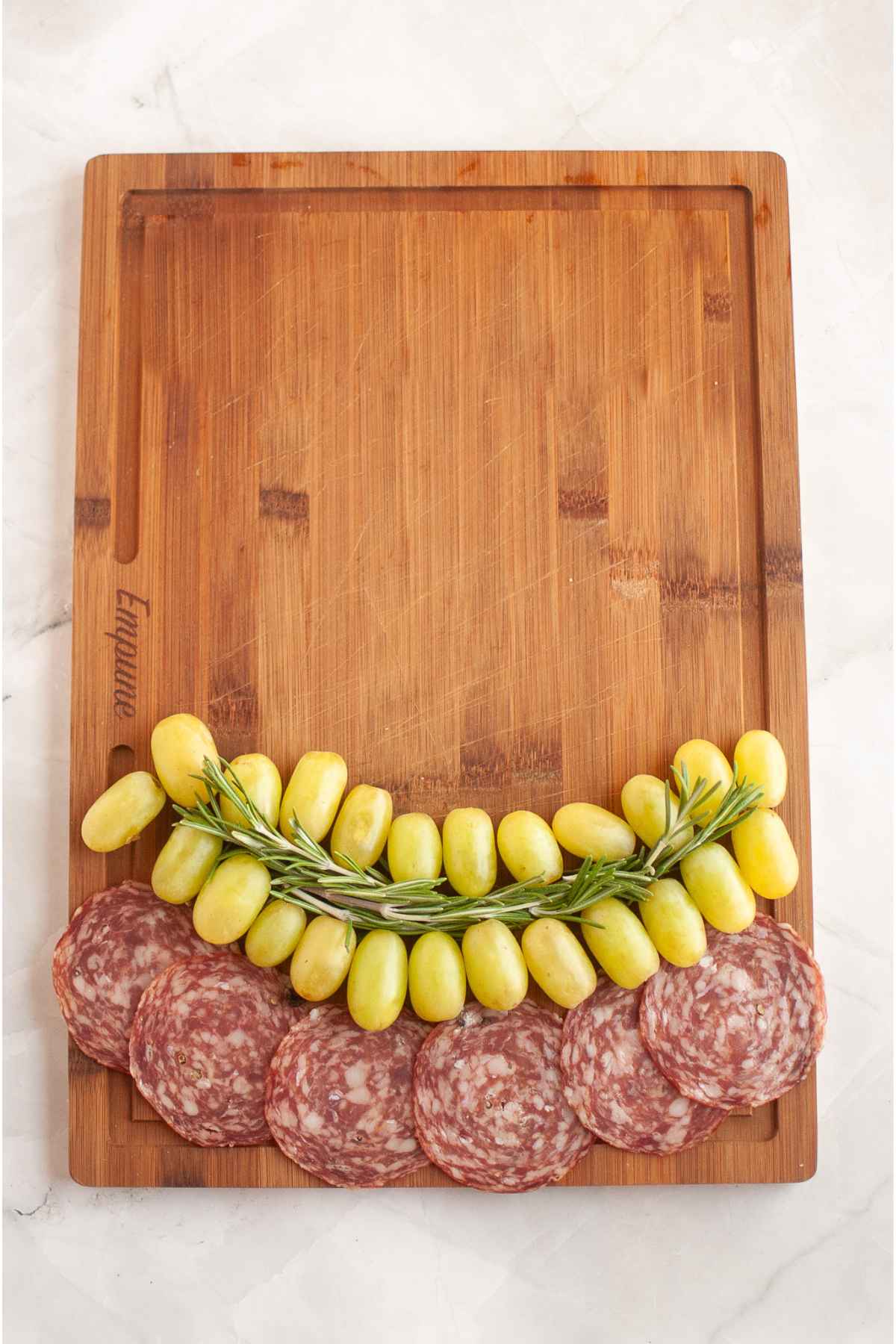 A wooden board with rows of green grapes, sprigs of rosemary, and slices of salami arranged at the bottom in preparation for assembling a holiday charcuterie board.
