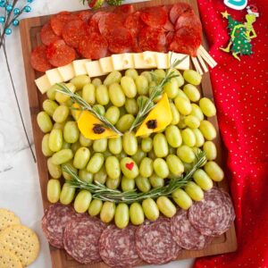 A completed Grinch charcuterie board featuring layers of pepperoni, cheese cubes, and green grapes shaped like the Grinch's face with rosemary eyebrows, cheese and raisin eyes, and a small red heart nose.
