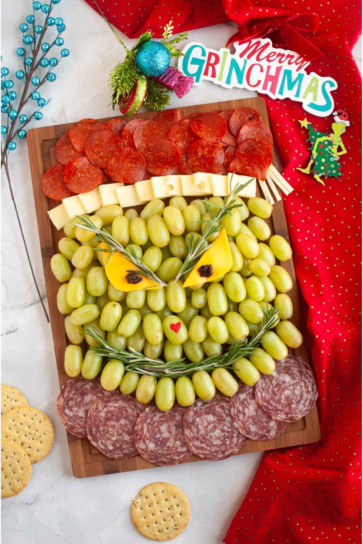 A completed Grinch charcuterie board featuring layers of pepperoni, cheese cubes, and green grapes shaped like the Grinch's face with rosemary eyebrows, cheese and raisin eyes, and a small red heart nose.