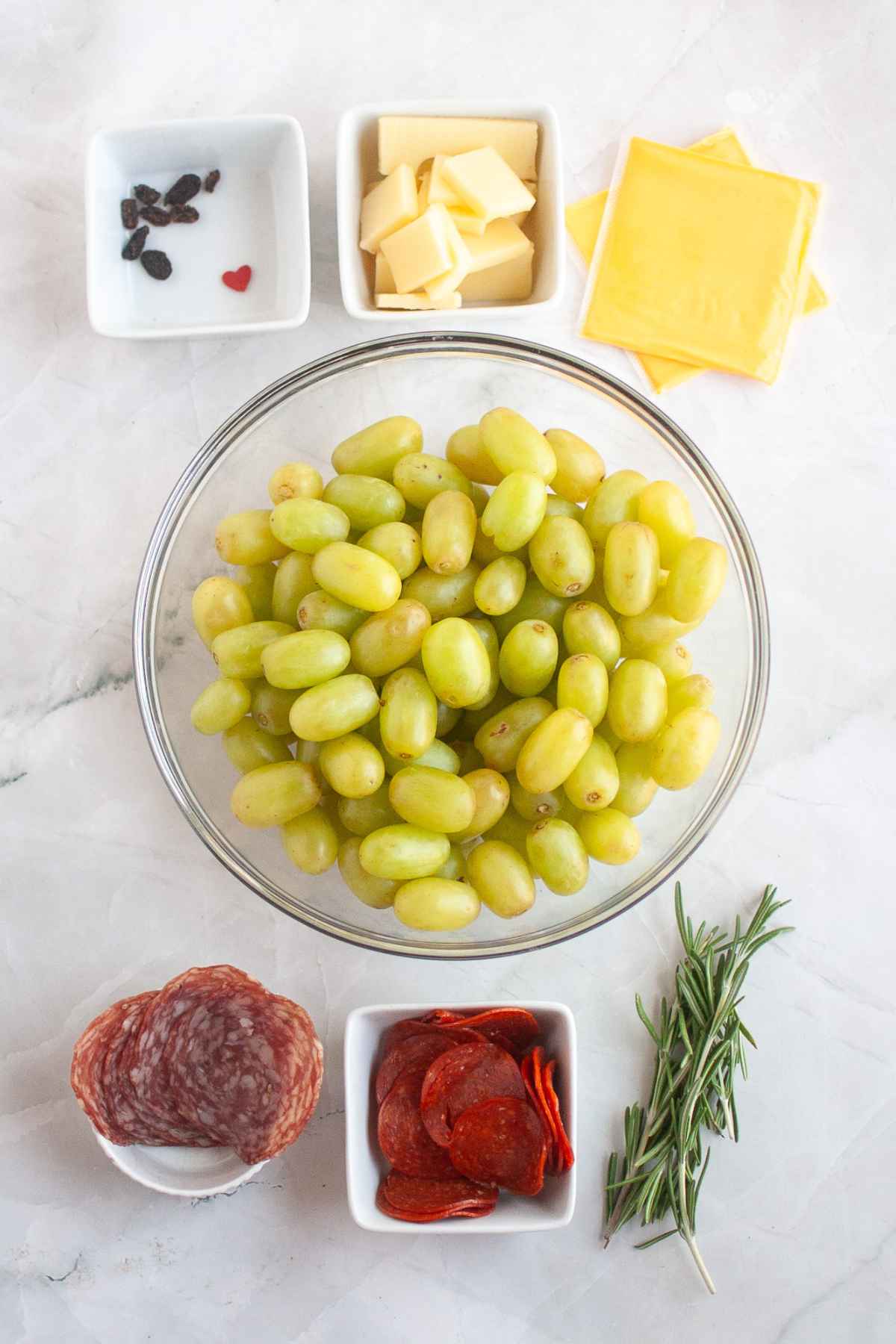A flat lay of ingredients for a charcuterie board, including green grapes, salami, pepperoni, cheese cubes, cheese slices, raisins, rosemary sprigs, and a small red candy heart.