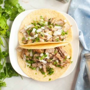 Two corn tortillas filled with shredded instant pot pork carnitas, copped onions, and cilantro on a white plate.