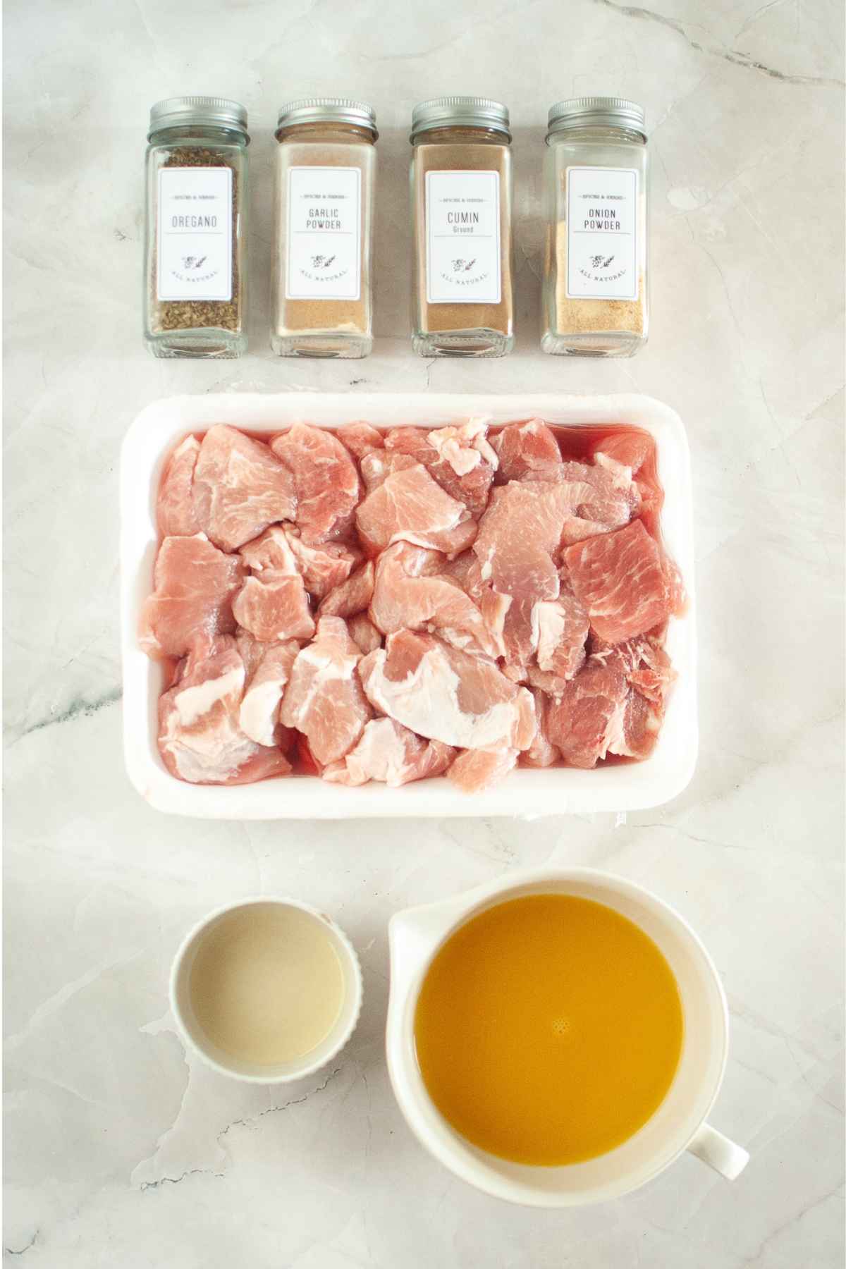 Overhead view of measured ingredients including raw pork chunks, broth, vinegar, and seasonings: oregano, garlic powder, cumin, and onion powder.