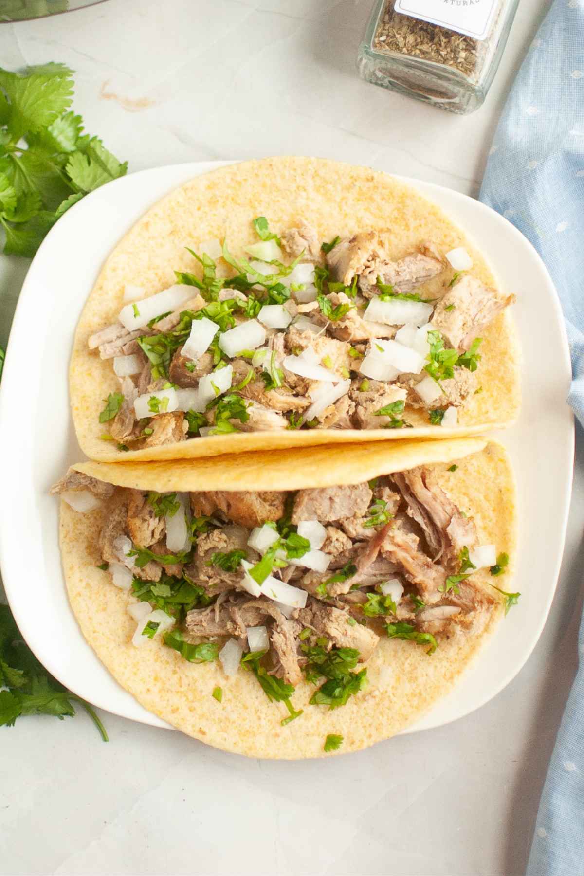 Two corn tortillas filled with shredded instant pot pork carnitas, copped onions, and cilantro on a white plate.