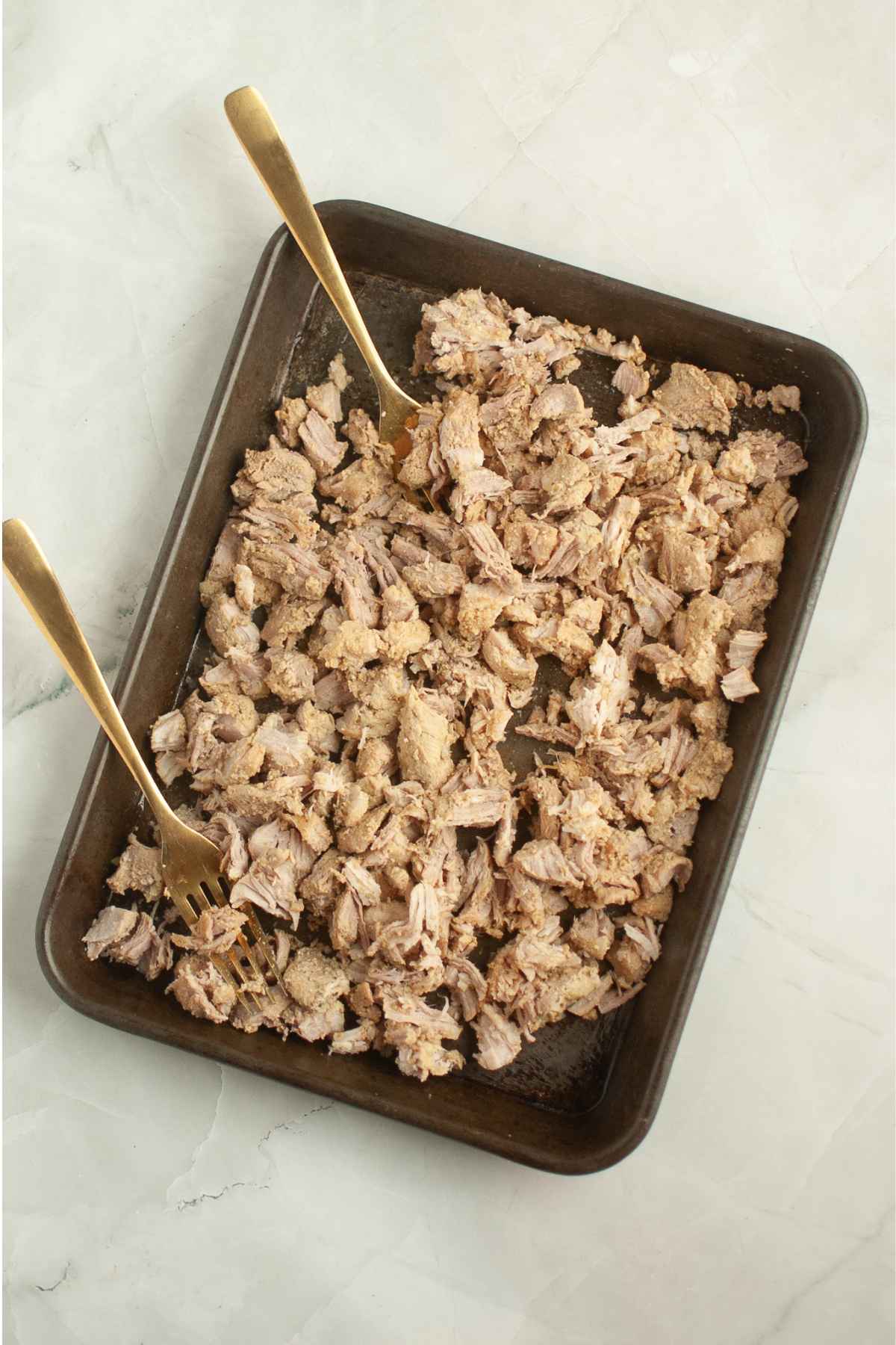 Shredded pork carnitas spread across a dark baking pan with two gold forks used for pulling the meat apart.