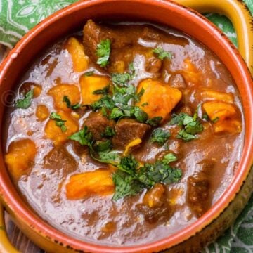 Close-up of stew in a rich red broth with chunks of meat and sweet potatoes topped with chopped cilantro.