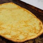 Baked keto pizza crust on a dark baking sheet, golden brown and slightly crisp around the edges.