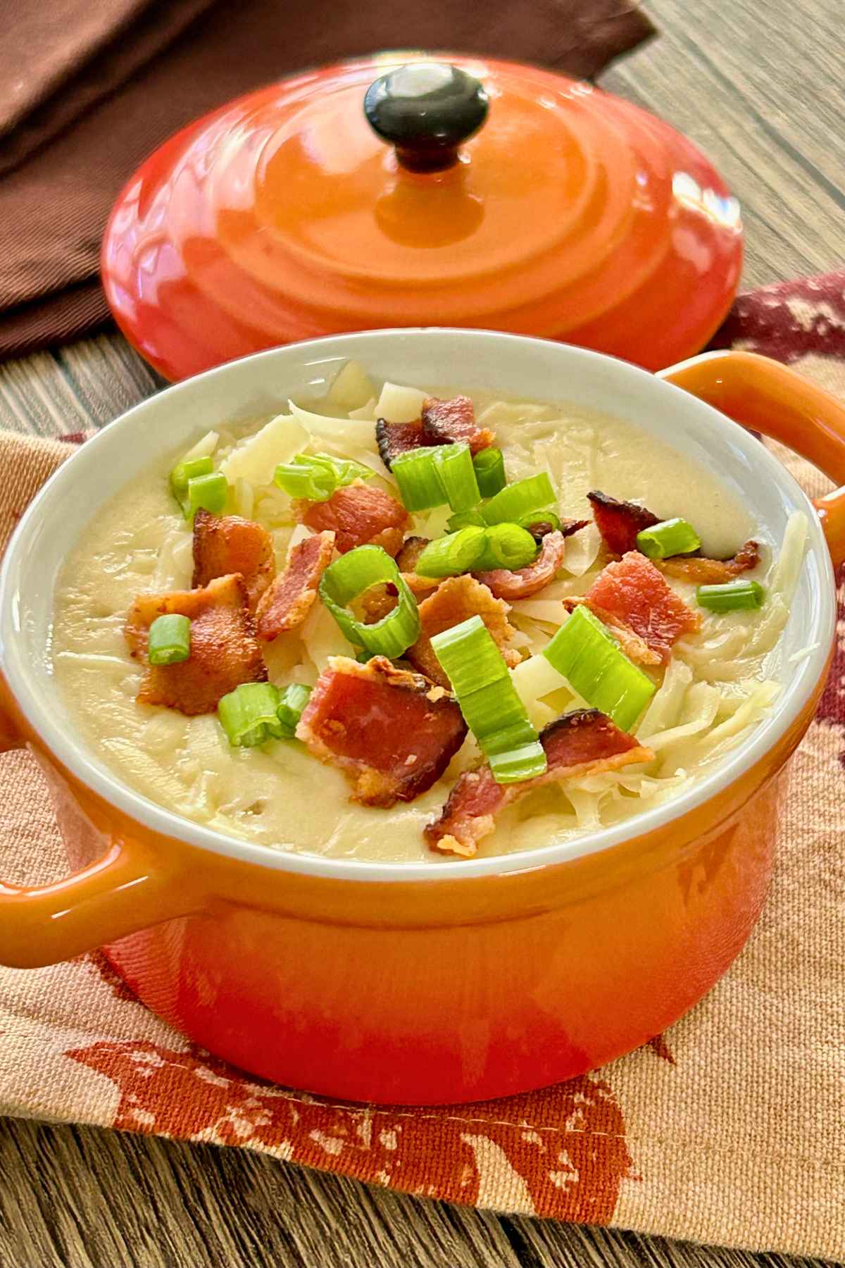 Creamy loaded potato soup served in an orange soup crock, topped with crispy bacon pieces, shredded cheese and sliced green onions.