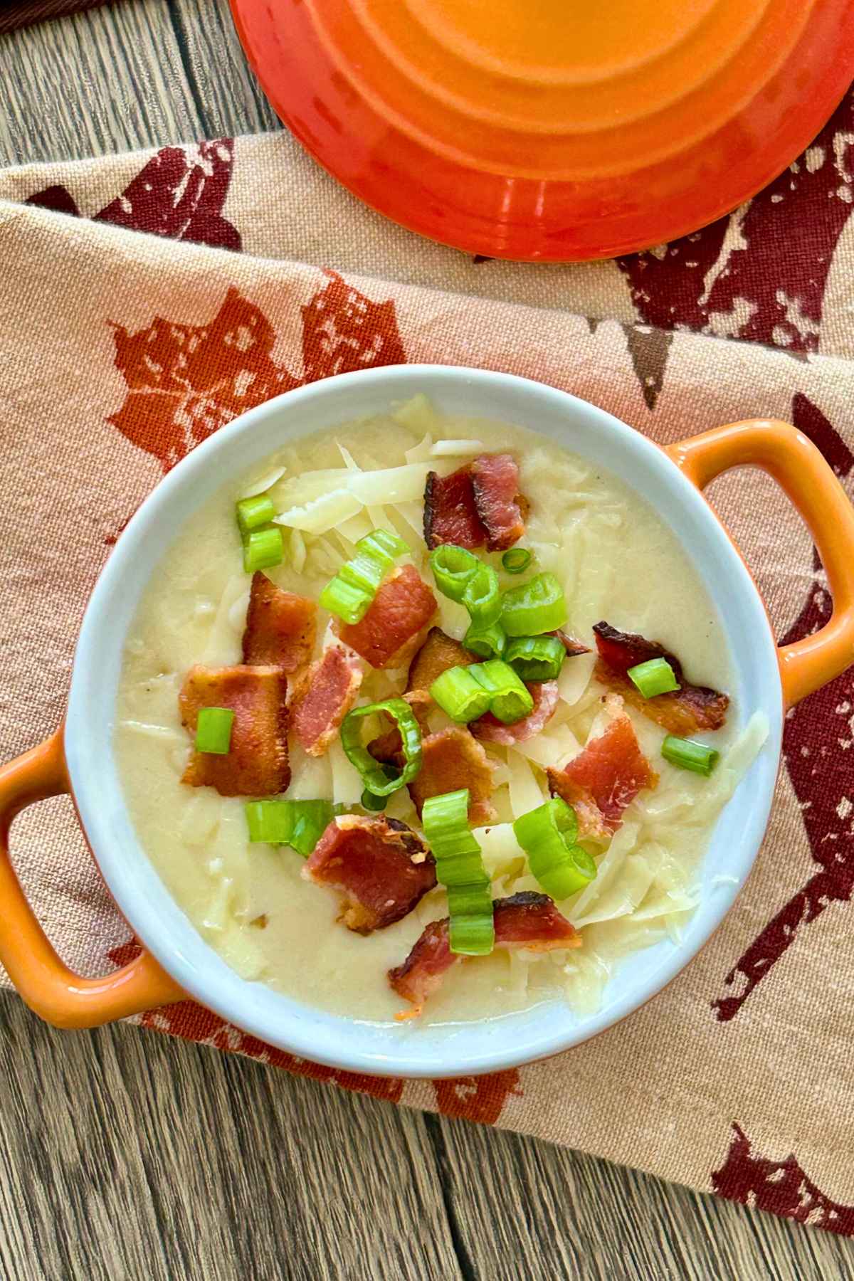Overhead image of bacon potato soup in an orange crock on a fall leaf printed napkin.