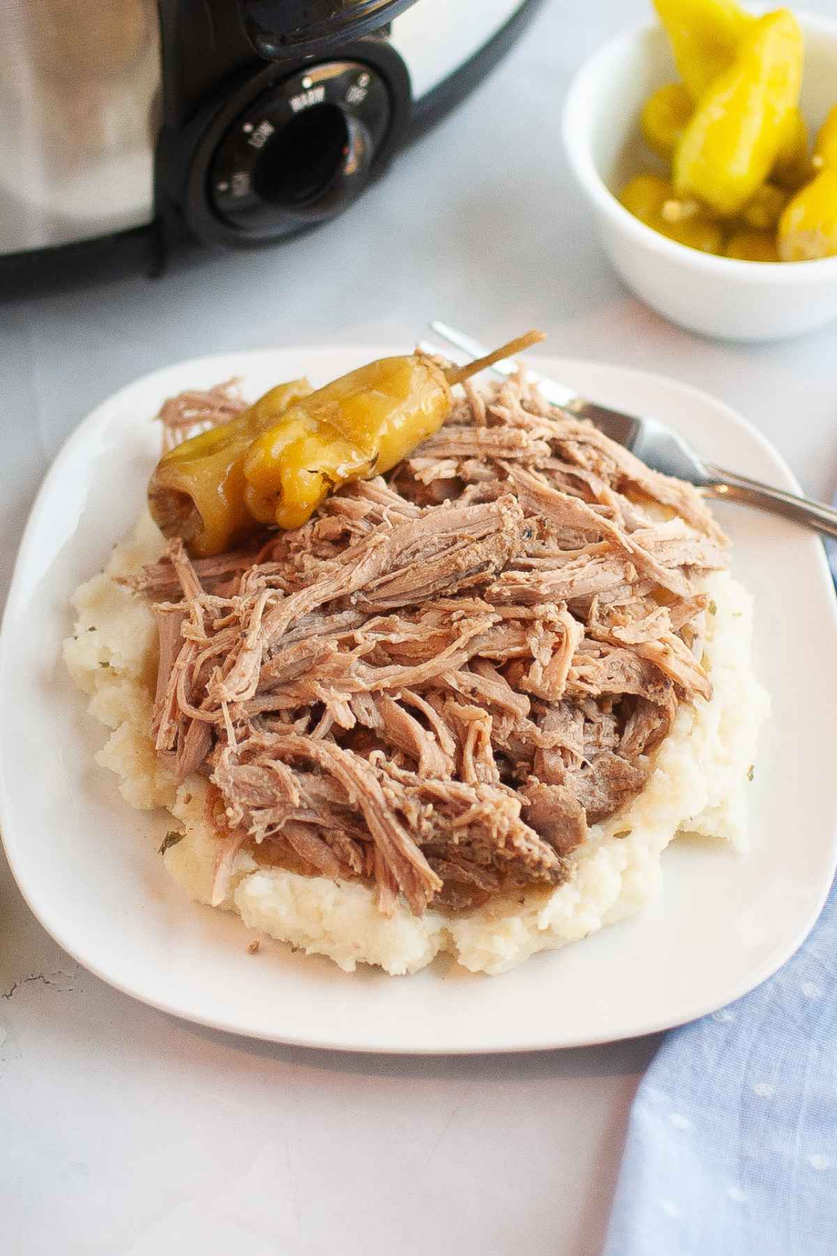 Shredded Mississippi Pork Roast served over mashed potatoes on a white plate topped with pepperoncini peppers beside a slow cooker