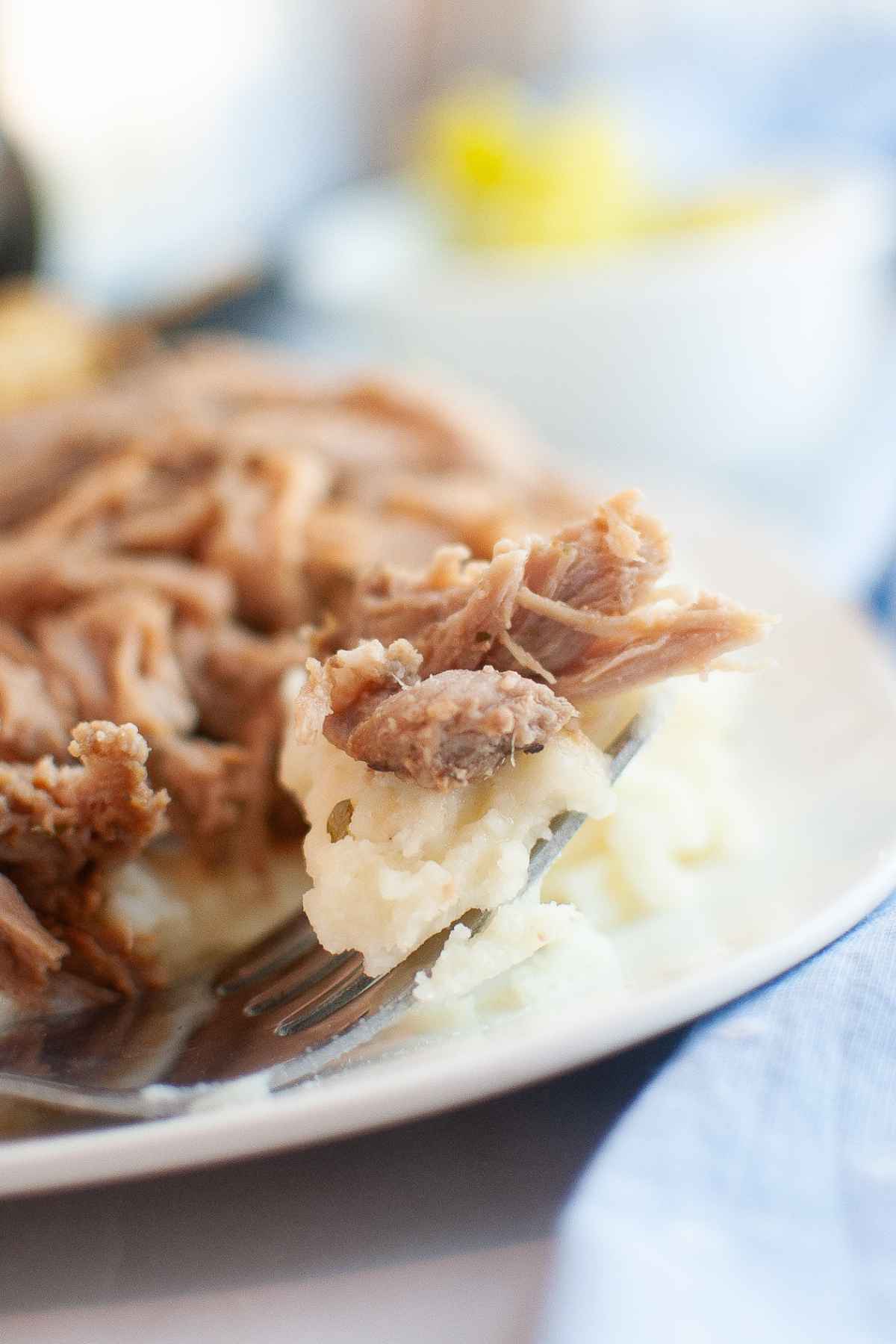 Close up of a fork holding a bite of shredded pork roast and mashed potatoes with the full plate blurred in the background.