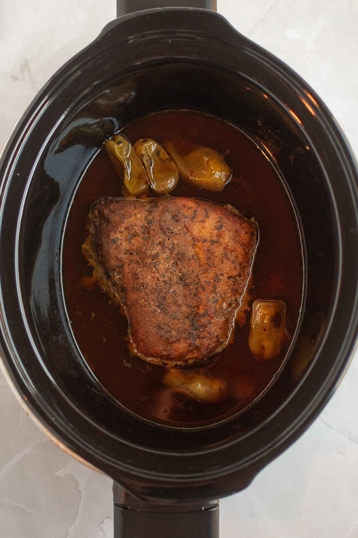Whole seasoned pork roast cooking in a slow cooker with pepperoncini pepers and rich brown juices.
