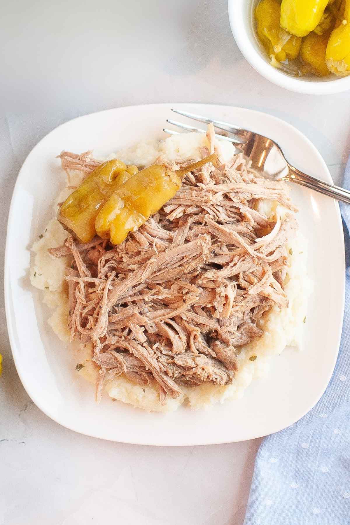 Overhead view of shredded Mississippi Pork Roast with pepperoncini peppers served on mashed potatoes next to a bowl of peppers.
