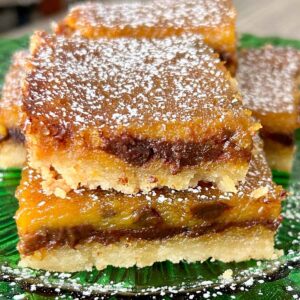 A close-up of pumpkin chocolate chip bars stacked on a green glass plate, dusted with powdered sugar, showing a rich golden-orange layer with melted chocolate throughout.