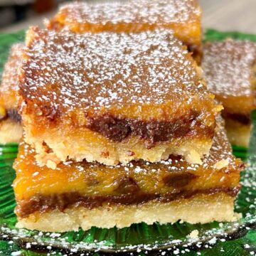 A close-up of pumpkin chocolate chip bars stacked on a green glass plate, dusted with powdered sugar, showing a rich golden-orange layer with melted chocolate throughout.