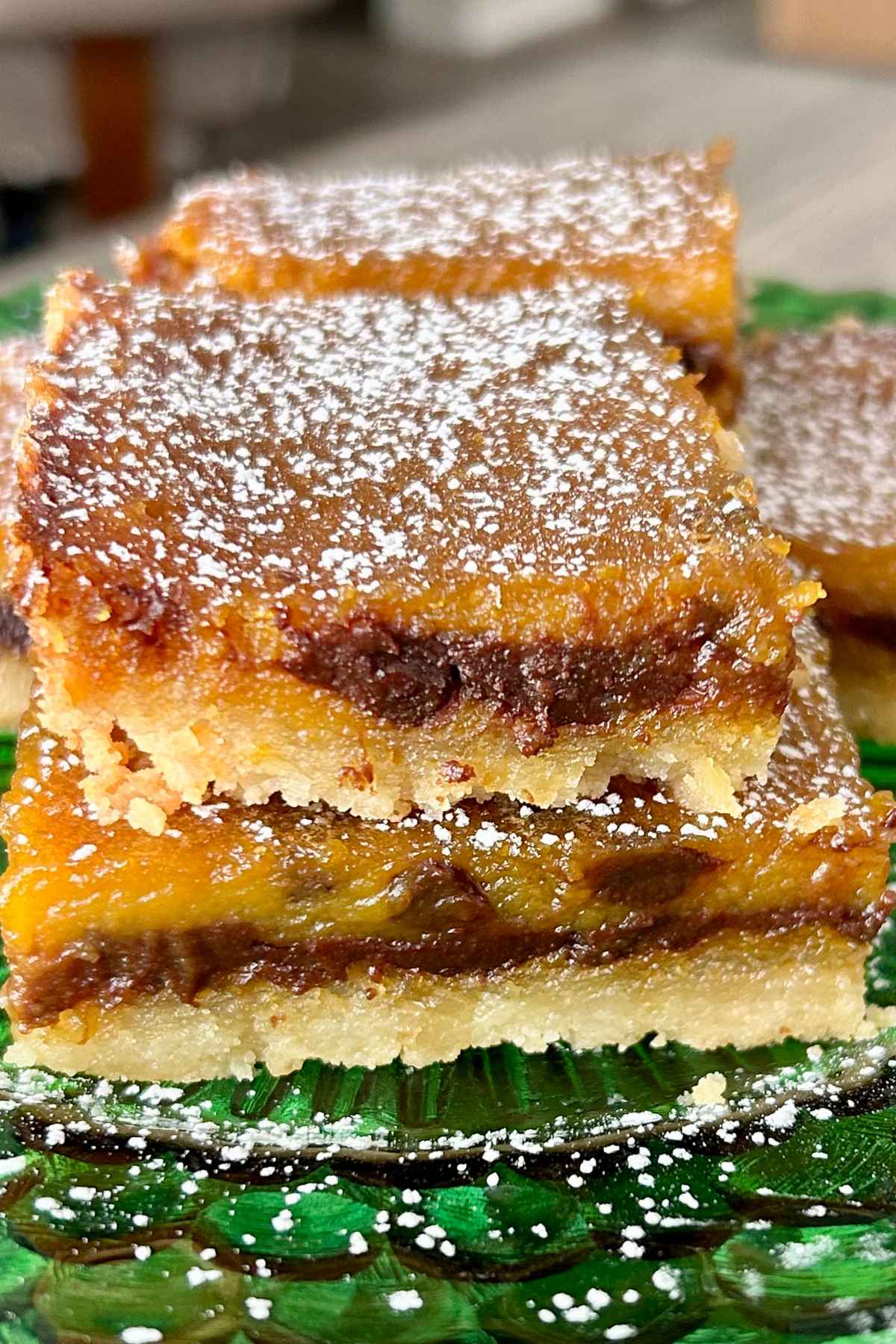 A close-up of pumpkin chocolate chip bars stacked on a green glass plate, dusted with powdered sugar, showing a rich golden-orange layer with melted chocolate throughout.
