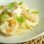 A plate of creamy shrimp linguine topped with sliced green onions, featuring large shrimp coated in a rich white sauce over thin pasta.