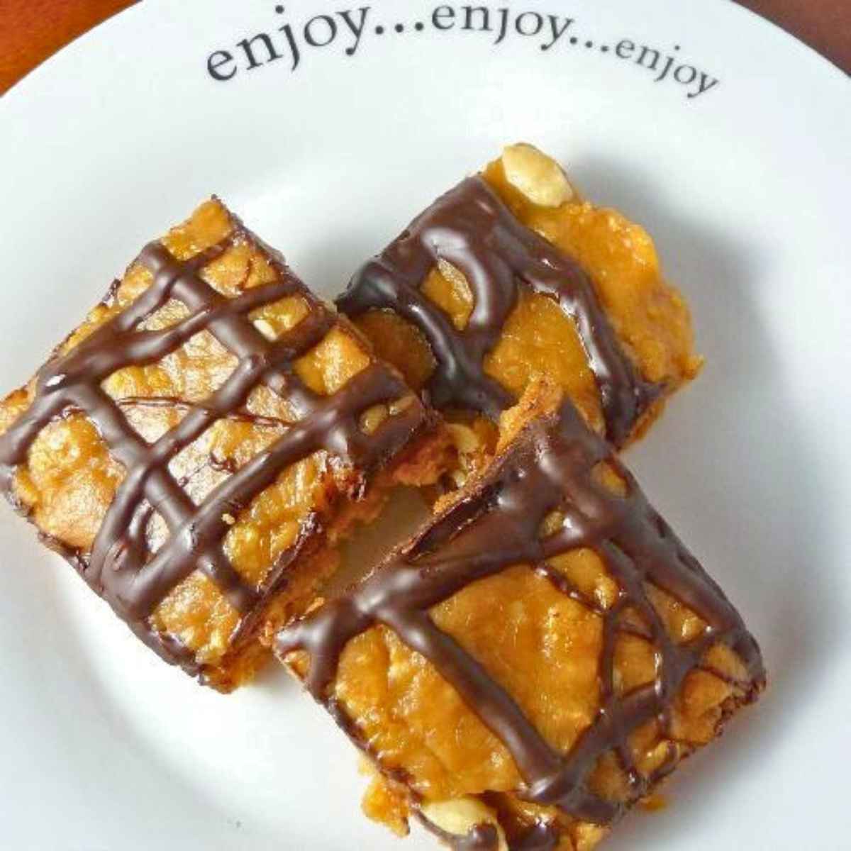 Three golden peanut butter cereal bars drizzled with chocolate, arranged on a white plate that reads "enjoy...enjoy...enjoy."