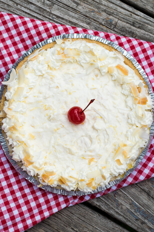 A whole Pina colada pie topped with whipped cream, toasted coconut flakes, and a single maraschino cherry in the center, set on a red checkered cloth.
