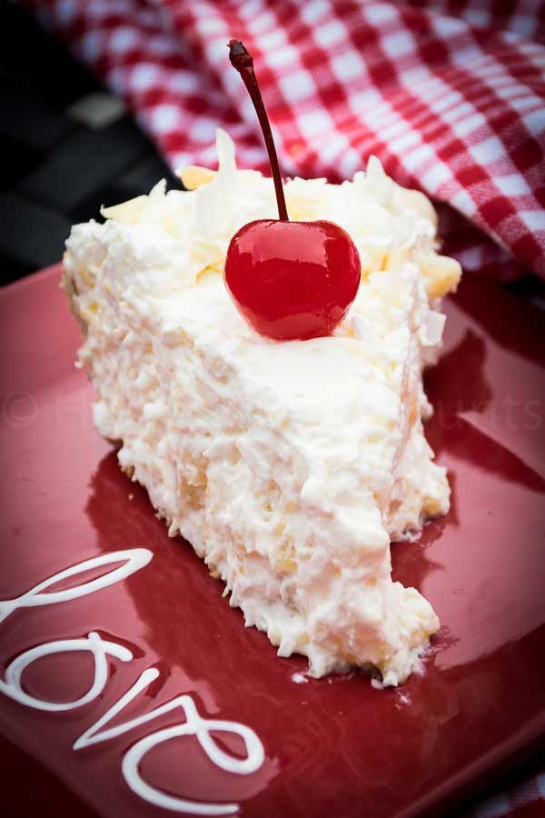A slice of creamy Pina Colada pie on a red plate with the word "love" garnished with a bright maraschino cherry and topped with whipped cream and coconut flakes.