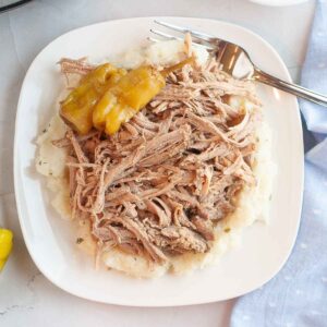 Overhead shot of shredded slow cooker Mississippi Pork Roast with pepperoncini peppers served over mashed potatoes on a white plate with a fork.